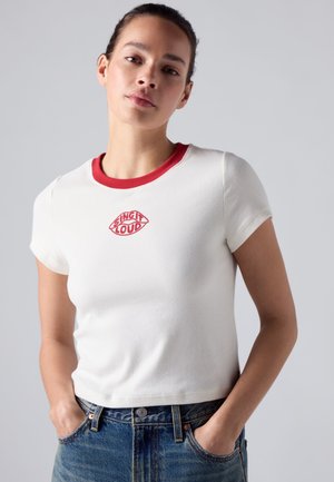 Jeune femme les mains dans les poches de son jean, portant un t-shirt court blanc avec un col rouge et le texte "Sing It Loud" à l'intérieur d'un motif en forme de lèvres.