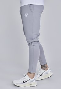 Lys grå slim-fit joggers lavet af et blødt stof, med et hvidt logo, sidelommer og ribbet talje, parret med hvide træningssko.