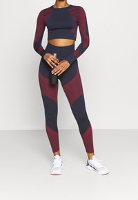 Marineblaues und burgunderfarbenes Sport-Set bestehend aus einem langärmeligen Cropped-Top und hochgeschnittenen Leggings mit strukturierten Paneelen, Wasserflasche in der Hand.