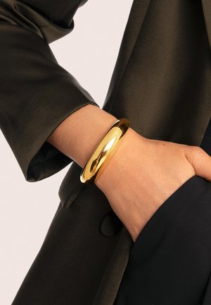 Pulsera de oro con una superficie lisa y pulida que se lleva en una muñeca, acompañada de una manga de tela oscura y texturizada.