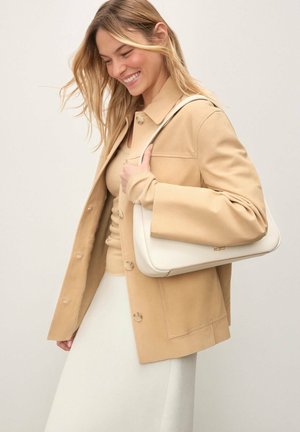 Femeie zâmbitoare cu părul blond lung poartă o jachetă bej peste un top asortat și o fustă albă, purtând o geantă de umăr albă.