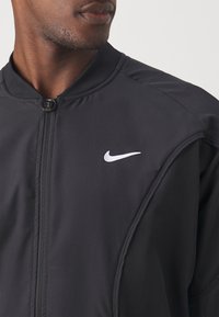 Mann trägt eine schwarze Nike-Trainingsjacke mit Reißverschluss, auf der Brust ist das weiße Nike-Swoosh-Logo zu sehen, während er seitlich blickt.
