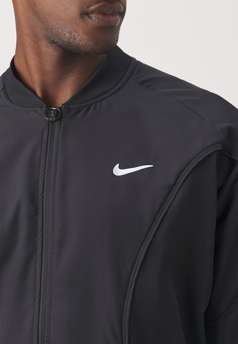 Man in een zwarte Nike zip-up jas, met het witte Nike swoosh logo op de borst, terwijl hij naar de zijkant kijkt.