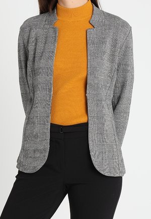 Frau trägt ein graues kariertes Sakko über einem senfgelben gerippten Rollkragenpullover und schwarzen Hosen, steht vor einem weißen Hintergrund.