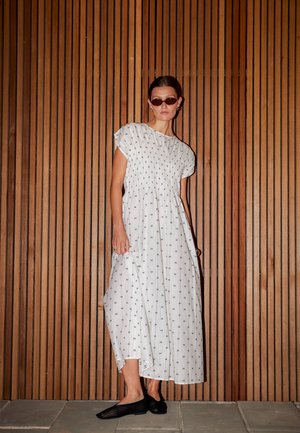Femme portant une robe midi blanche à motifs et des ballerines noires en maille, debout devant un fond de panneaux en bois vertical, portant des lunettes de soleil foncées.