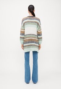 Πλεκτό πουλόβερ με πολύχρωμες ρίγες σε μέντα, κρέμα, καφέ και γκρι. Έχει μακριά μανίκια και φαρδιά εφαρμογή, με πιο μακρύ πίσω στρίφωμα. Φαίνονται τζιν από κάτω.