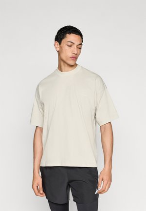 Beżowy t-shirt z krótkim rękawem o luźnym kroju, z okrągłym dekoltem i miękką fakturą. Bez widocznych wzorów; noszony z czarnymi szortami.