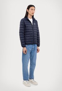 Navy puffer jacket with a quilted design, high collar, and front zipper. Paired with blue denim jeans and light sneakers.