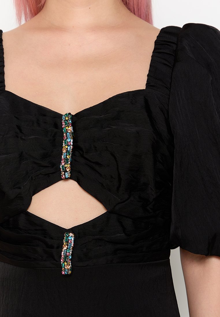 Frau trägt ein schwarzes Kleid mit Puffärmeln, Ausschnitt in der Mitte und zwei vertikalen, mehrfarbigen Edelsteindekorationen an der Vorderseite.