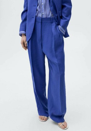 Helle blaue Leinen-Hosen mit weitem Bein und hohem Bund, ausgestattet mit Seitentaschen und einem bequemen Schnitt. Kombiniert mit einem gestreiften Hemd und Sandalen.