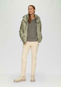 Licht olijfgroene pufferjack met capuchon, grijze gebreide trui, crèmekleurige broek en beige enkellaarsjes. Soepele texturen en casual pasvorm.