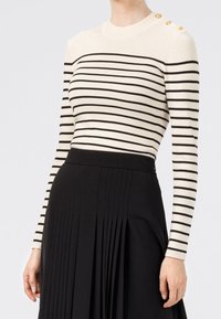 Femme portant un pull crème à manches longues avec des rayures horizontales noires et des boutons dorés sur l'épaule, associé à une jupe plissée noire.