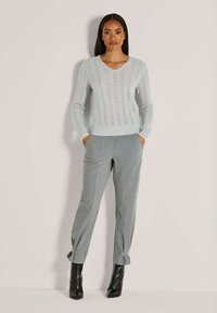 Frau in einem hellgrauen gemusterten Pullover, grauen maßgeschneiderten Hosen mit Knöchelbändern und schwarzen Absatzstiefeln, die mit den Händen in den Taschen steht.