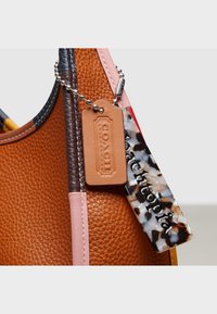 Läheltä kuvattu ruskea teksturoitu nahkalaukku vaaleanpunaisilla ja tummanruskeilla reunoilla, jossa on vaaleanruskea Coach-tägi ja marmoroitu avaimenperi, jossa lukee "Coach x Georgia."