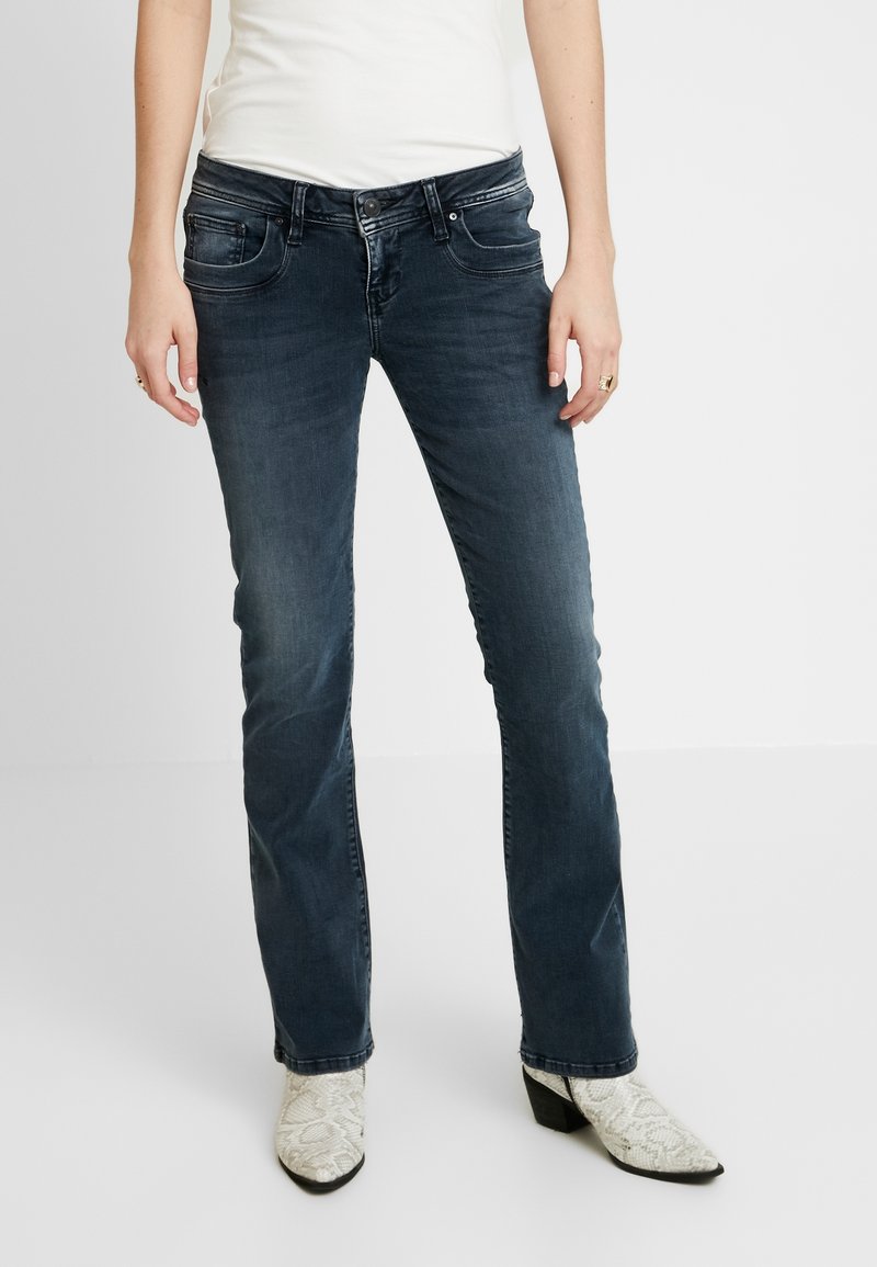 Jean en denim bleu foncé à coupe slim, avec poches avant, fermeture par boutons et légère décoloration. Associé à des bottines blanches à motifs.