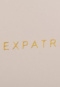 Helles beiges Textur-Hintergrund mit geprägten Buchstaben, die "EXPAT" in Gold schreiben. Sauberes Design, moderne Typografie, minimalistische Stil.