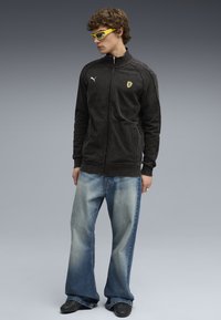 Sort lynlåsjakke med logo, parret med lyseblå flagrende jeans. Modellen bærer gule solbriller og sorte sko. Glat tekstur generelt.