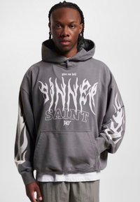 Grå oversized hettegenser med frontlomme. Har fet hvit tekst "SINNER" og "SAINT," samt flammemønster på ermnene. Teksturert stoff.