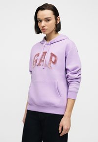 Νεαρή γυναίκα με κοντά σκούρα μαλλιά που φοράει ένα ανοιχτό μωβ φούτερ GAP και μαύρο παντελόνι, στέκεται μπροστά σε ένα απλό λευκό φόντο.