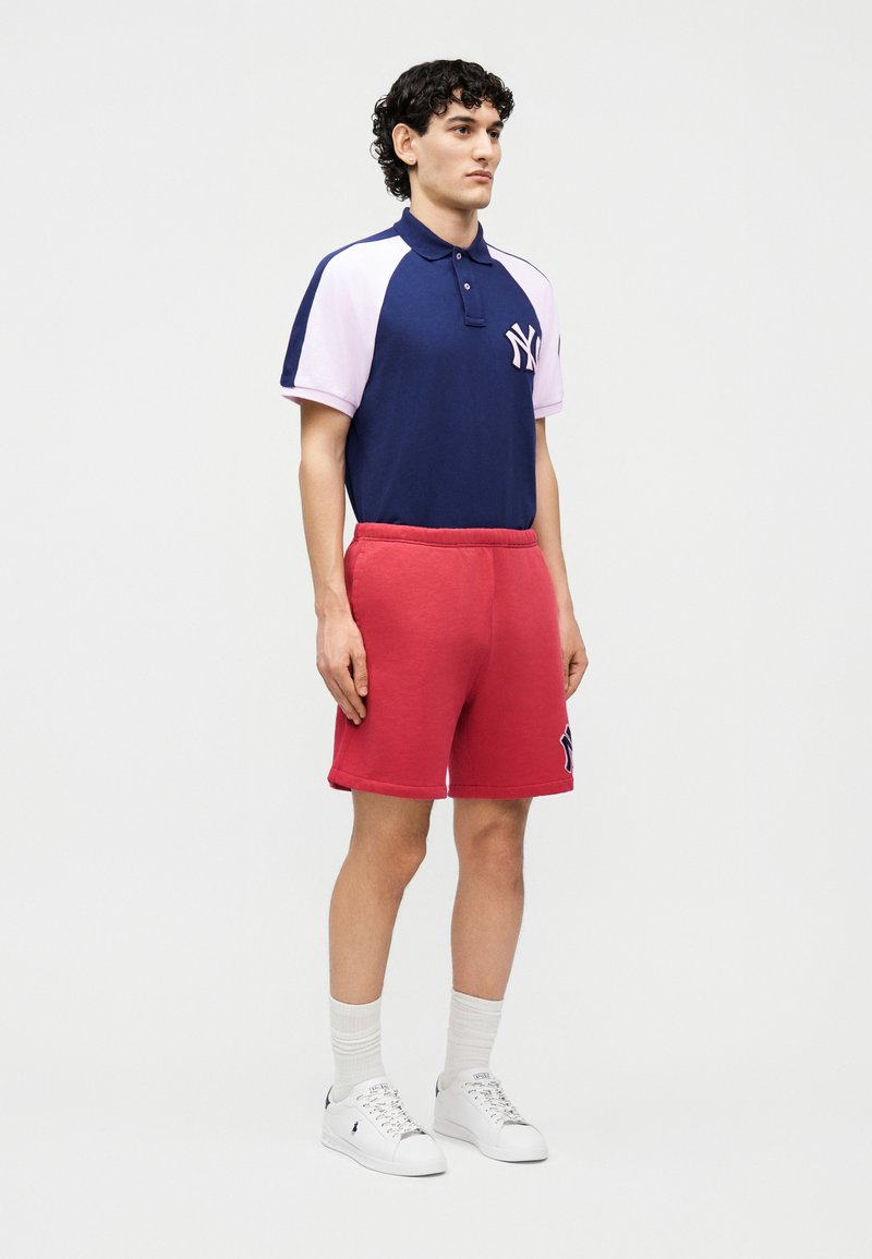 Jonge man staand in rode shorts, een marineblauw en roze poloshirt met NY-logo, witte sokken en witte sneakers tegen een witte achtergrond.