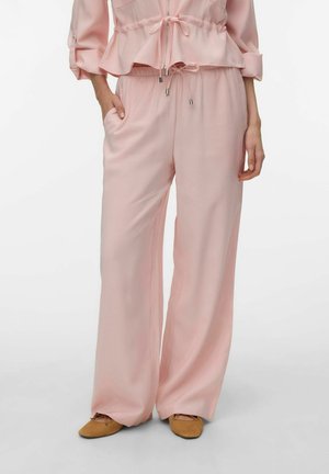 Kobieta ubrana w jasnoróżowe szerokie spodnie z regulacją w pasie na sznurek oraz dopasowany długi rękaw, w połączeniu z beżowymi balerinami.