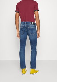 Blå denimjeans med slim fit, signatur bakfickor och en svart logobälte, kombinerat med gula högkängor.