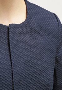 Granatowy teksturowany materiał z geometrycznym wzorem. Projekt ma zaokrąglony dekolt i prostą, minimalistyczną zapinkę.