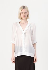 Blusa leggera bianca con maniche a sbuffo, scollatura a V e rifiniture in pizzo decorative lungo il davanti. Tessuto morbido con un fit rilassato.