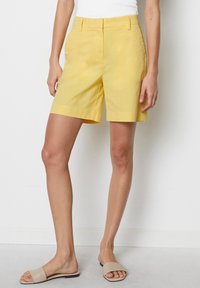 Vrouw in hooggeplaatste gele knielange shorts, witte top en beige platte slidesandalen die tegen een witte muur staat.