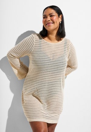 Smiling woman with shoulder-length black hair wearing a beige crochet-style long-sleeve dress, standing with hands behind her back.