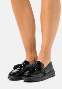 Mocassins en cuir verni noir avec un bout arrondi, des détails à pampilles et une épaisse semelle en caoutchouc noir. Texture lisse et design élégant.