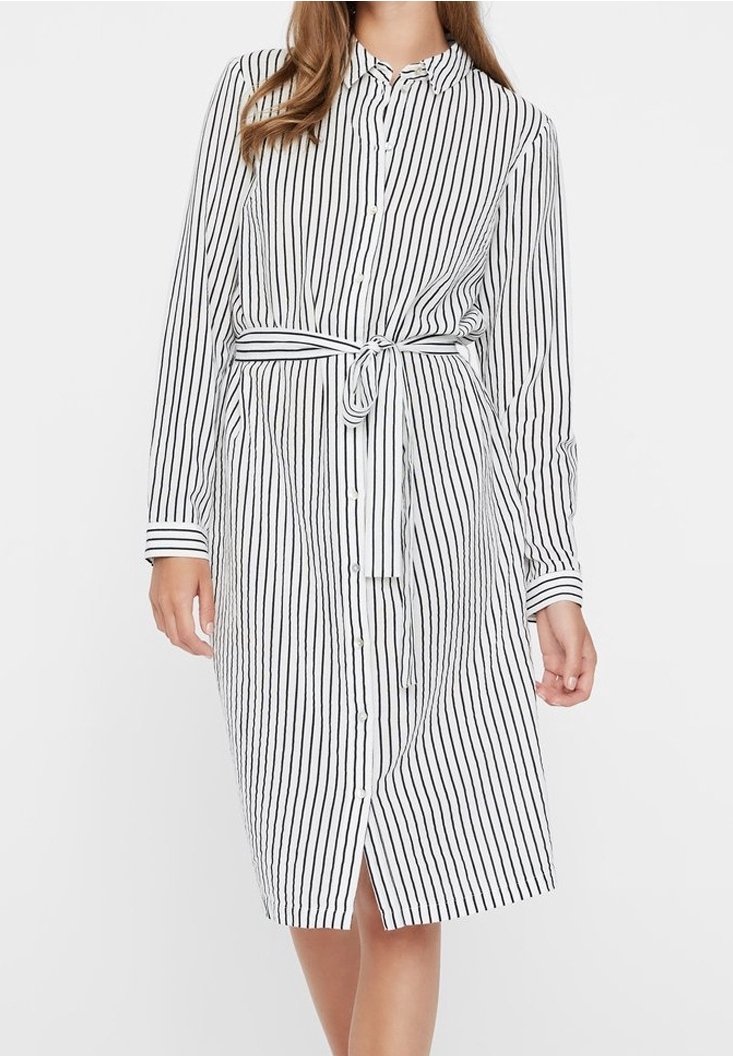 Femme portant une robe-chemise blanche mi-longue aux rayures verticales noires, boutonnée sur le devant, manches longues et ceinture nouée à la taille.