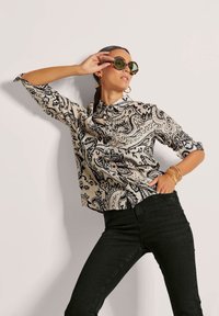 Schwarz-weißes Bluse mit Paisleymuster und Kragen, hochgekrempelte Ärmel; kombiniert mit schwarzen Jeans. Modell trägt übergroße runde Sonnenbrillen.