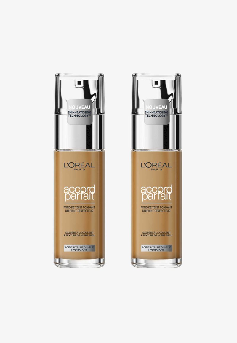 Deux flacons de fond de teint L'Oréal Paris, chacun avec un corps en verre transparent, une pompe en acier inoxydable et un liquide de couleur beige à l'intérieur. Les étiquettes décrivent les caractéristiques du produit.