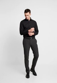 Czarna koszula zapinana na guziki, dopasowana, w połączeniu z ciemnoszarymi spodniami. Akcesoria to czarny pasek i wypolerowane czarne buty. Gładkie tło.