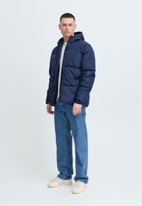 Jachetă puffer albastră închis cu glugă, cu un design matlasat și închidere cu fermoar, asortată cu blugi albastri deschis și adidași bej.