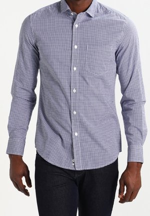 Mann trägt ein langärmeliges, blau-weiß kariertes Hemd mit Knöpfen und dunkle Hose, steht vor einem schlichten weißen Hintergrund.