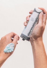 Dermalogica Hydro Masque Exfoliant in einer Tube mit Quetschverschluss, graues und weißes Design, auf einer Hand aufgetragen. Das Produkt ist hellblau und hat eine strukturierte Oberfläche.