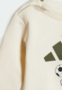 Cream-colored sweatshirt with a textured finish, featuring green geometric logos and a soccer ball graphic on the front. Includes snaps at the shoulder.