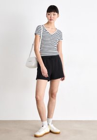 Svart shorts med elastisk midje i kombinasjon med en svart og hvit stripete v-hals skjorte. Hvite joggesko med gule detaljer og en crossbody veske.