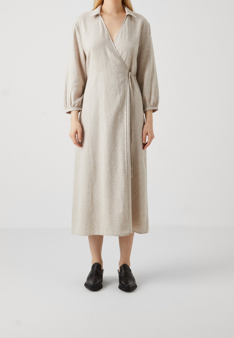 Robe portefeuille en lin beige avec un col en V, des manches longues bouffantes et un détail noué à la taille, associée à des chaussures noires. Tissu simple et texturé.