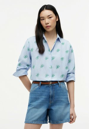 Junge Frau mit langen dunklen Haaren trägt ein blau gestreiftes Hemd mit grünen Initialen, braunen Gürtel und Jeansshorts vor einem schlichten weißen Hintergrund.