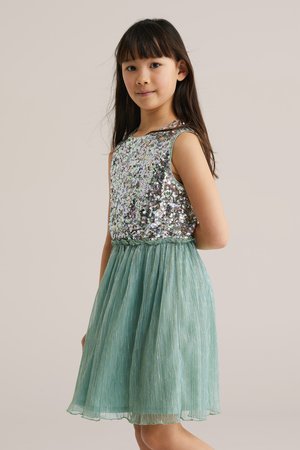 Fată tânără stând în profil, purtând o rochie fără mâneci cu un top argintiu din paiete și o fustă plisată verde deschis.