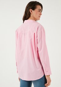 Pink button-up shirt with long sleeves, back pleat, and rounded hem. Made from smooth, lightweight fabric. Features cuffs with button details.