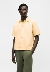 Άνδρας που φοράει ανοιχτό κίτρινο πουκάμισο με κοντά μανίκια και τσέπη στο στήθος, μαζί με χακί παντελόνι, στέκεται με το ένα χέρι στην τσέπη.