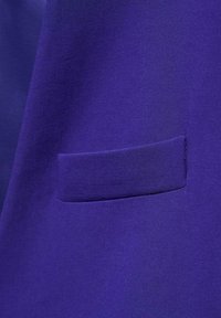 Fioletowa tkanina o gładkiej fakturze ma prostokątną nakładkę kieszeni. Materiał wydaje się lekki i dobrze skrojony.