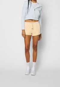 En person står iförd en ljusblå kort hoodie och pastellorange shorts, i kombination med vita sneakers och ankelsockor.