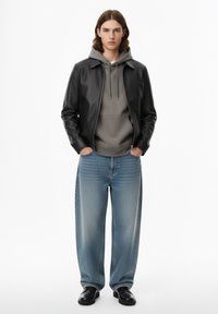 Giacca di pelle nera, felpa grigia con cappuccio e jeans a gamba larga azzurro chiaro. Texture lisce, colori neutri e vestibilità rilassata. Scarpe nere completano il look.