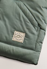 Tissu matelassé vert avec poche cousue et étiquette beige montrant un caméléon et le texte "PLAY ALL DAY GO EXPLORE."