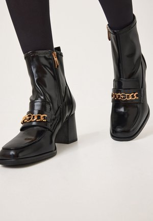 Botas de tobillo negras de cuero brillante con un tacón grueso, cierre de cremallera y un acento de cadena dorada a lo largo de la correa frontal.